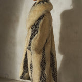 A model in profile wearing a long, voluminous beige faux fur coat with bold black patterned panels, a black wool cap, and black heels.