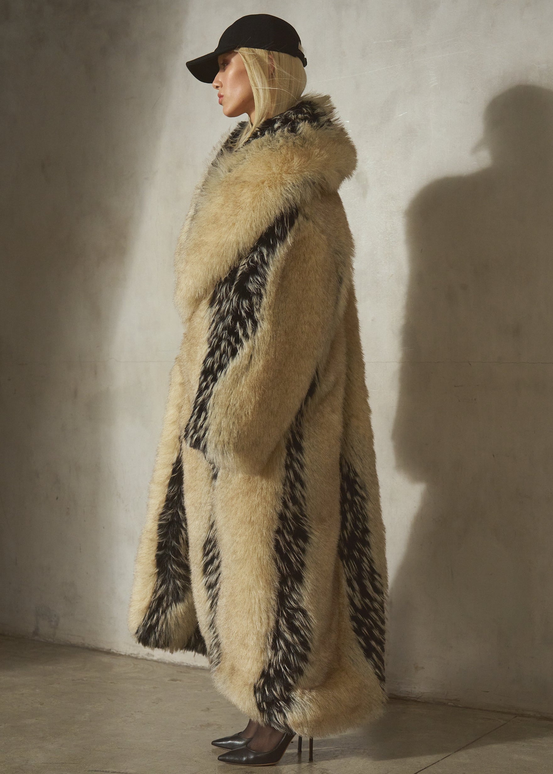 A model in profile wearing a long, voluminous beige faux fur coat with bold black patterned panels, a black wool cap, and black heels.