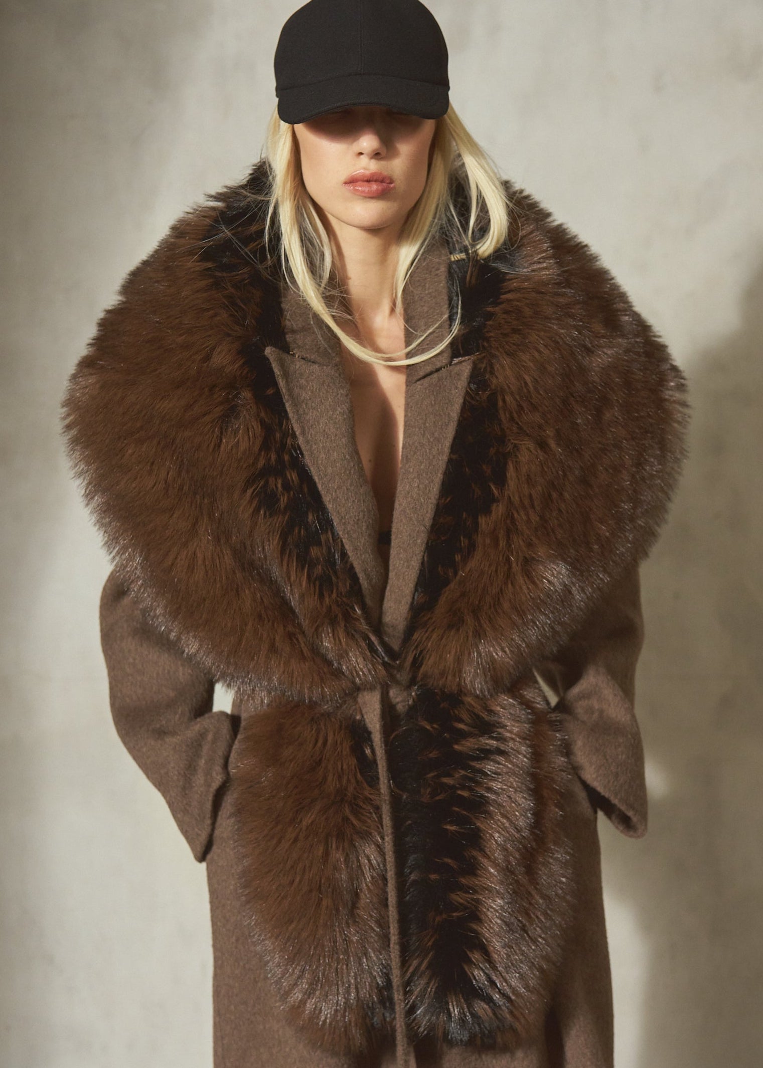 A model wearing a long brown faux fur collar over a wool coat, paired with a black baseball cap, standing against a textured concrete wall.