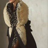 A woman wearing a long black coat with a large beige and black fur collar, a black hat, and sheer black tights, standing against a textured concrete wall.