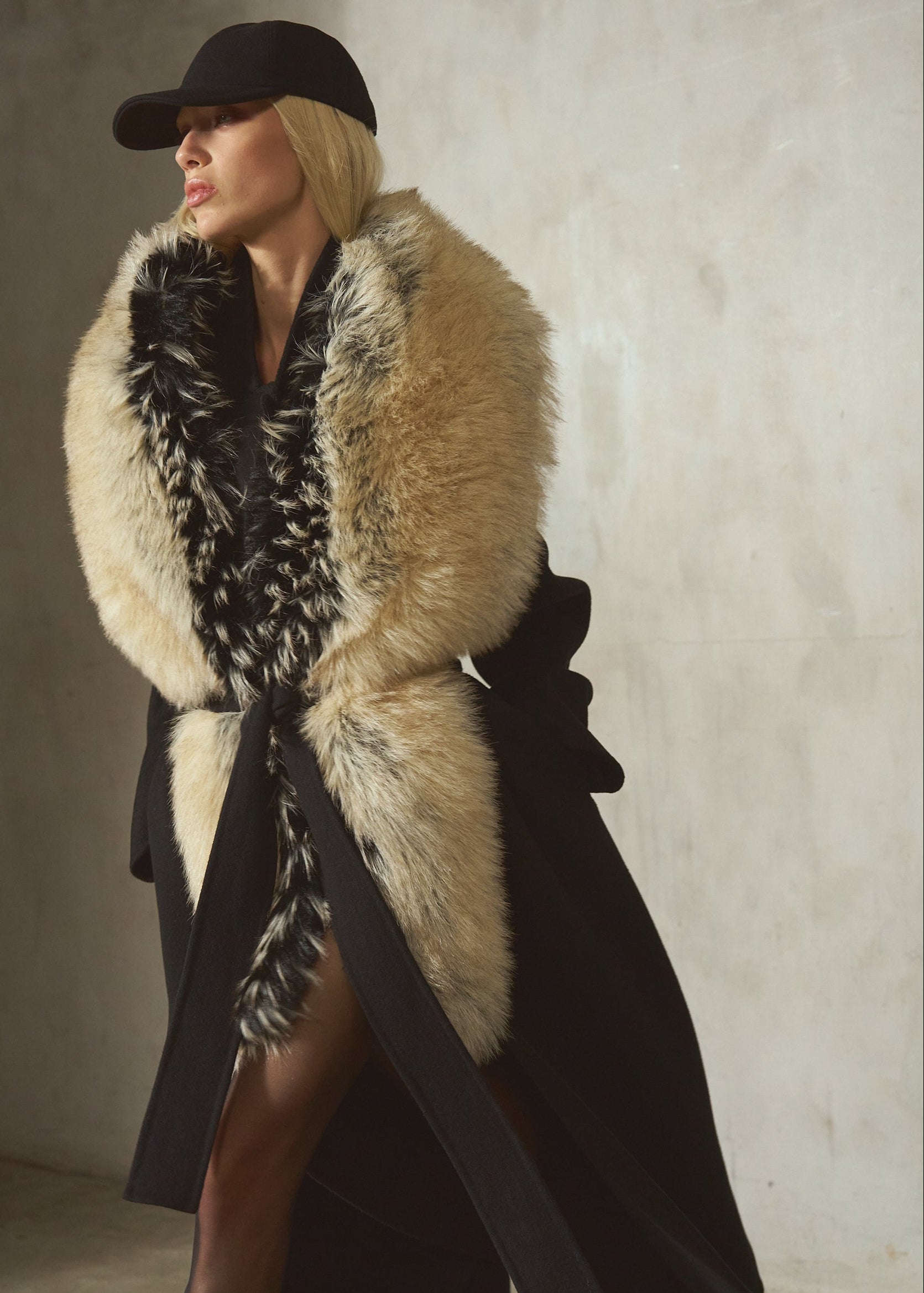 A woman wearing a long black coat with a large beige and black fur collar, a black hat, and sheer black tights, standing against a textured concrete wall.