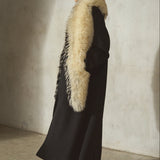 A model in profile wearing a long black coat with a large cream/black-colored faux fur collar, a black baseball cap, and black high heels.