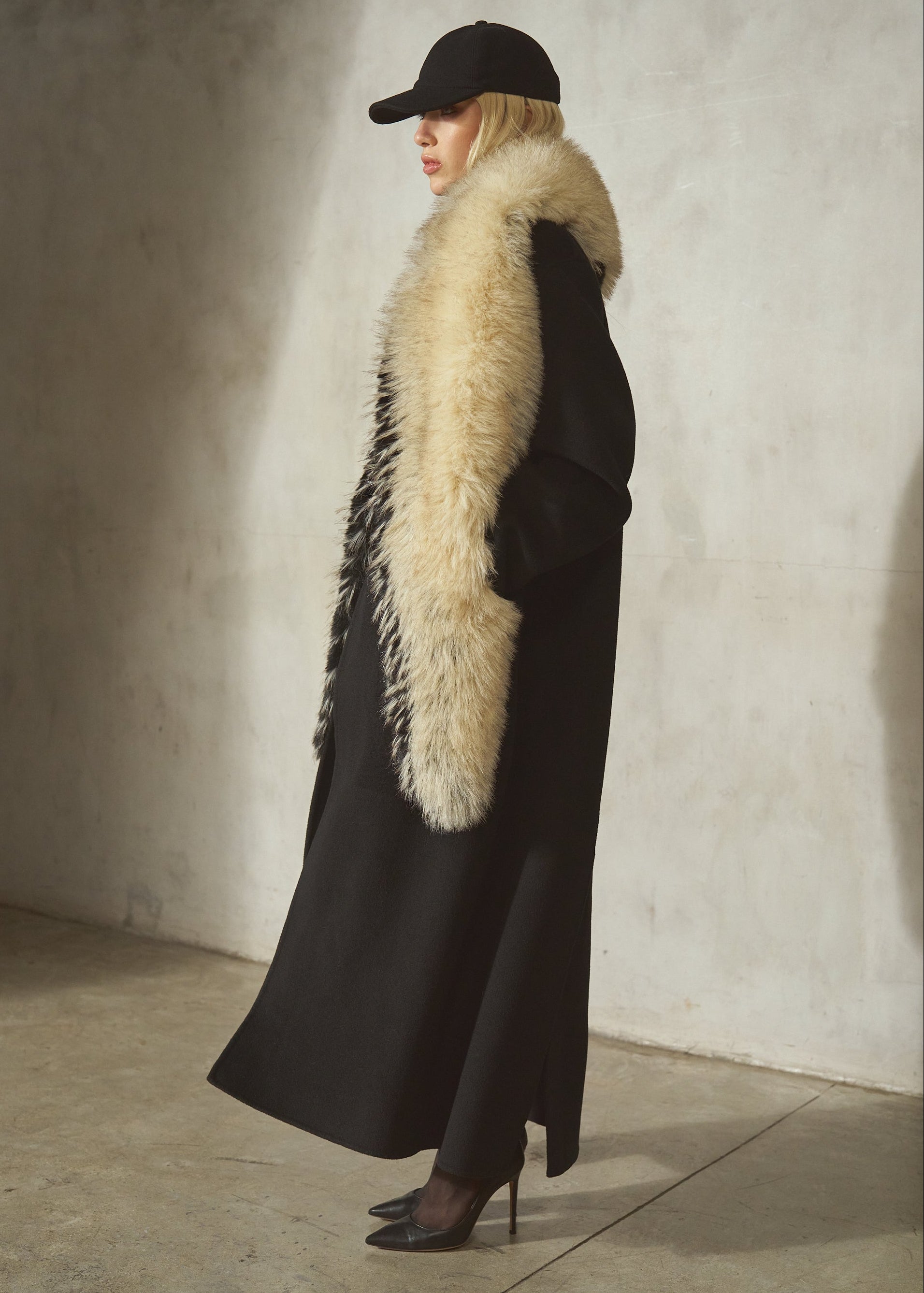A model in profile wearing a long black coat with a large cream/black-colored faux fur collar, a black baseball cap, and black high heels.
