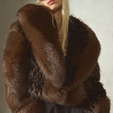 A model wearing a luxurious dark brown fur jacket with a wide collar, a black cap, and black trousers, posed against a textured gray wall.