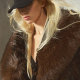 A close-up lifestyle shot of a model wearing a luxurious dark brown black faux fur coat and a black baseball cap, with her blonde hair blowing in the wind against a neutral gray background.