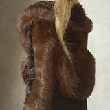 A woman shown from the back wearing a luxurious dark brown black fur coat with a large shawl collar, a black baseball cap, and black pants, standing against a textured concrete wall.