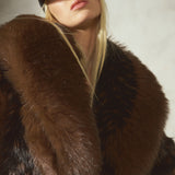 A close-up of a model wearing a luxurious dark brown black fur coat with a wide collar and a black cap, set against a textured light gray background.