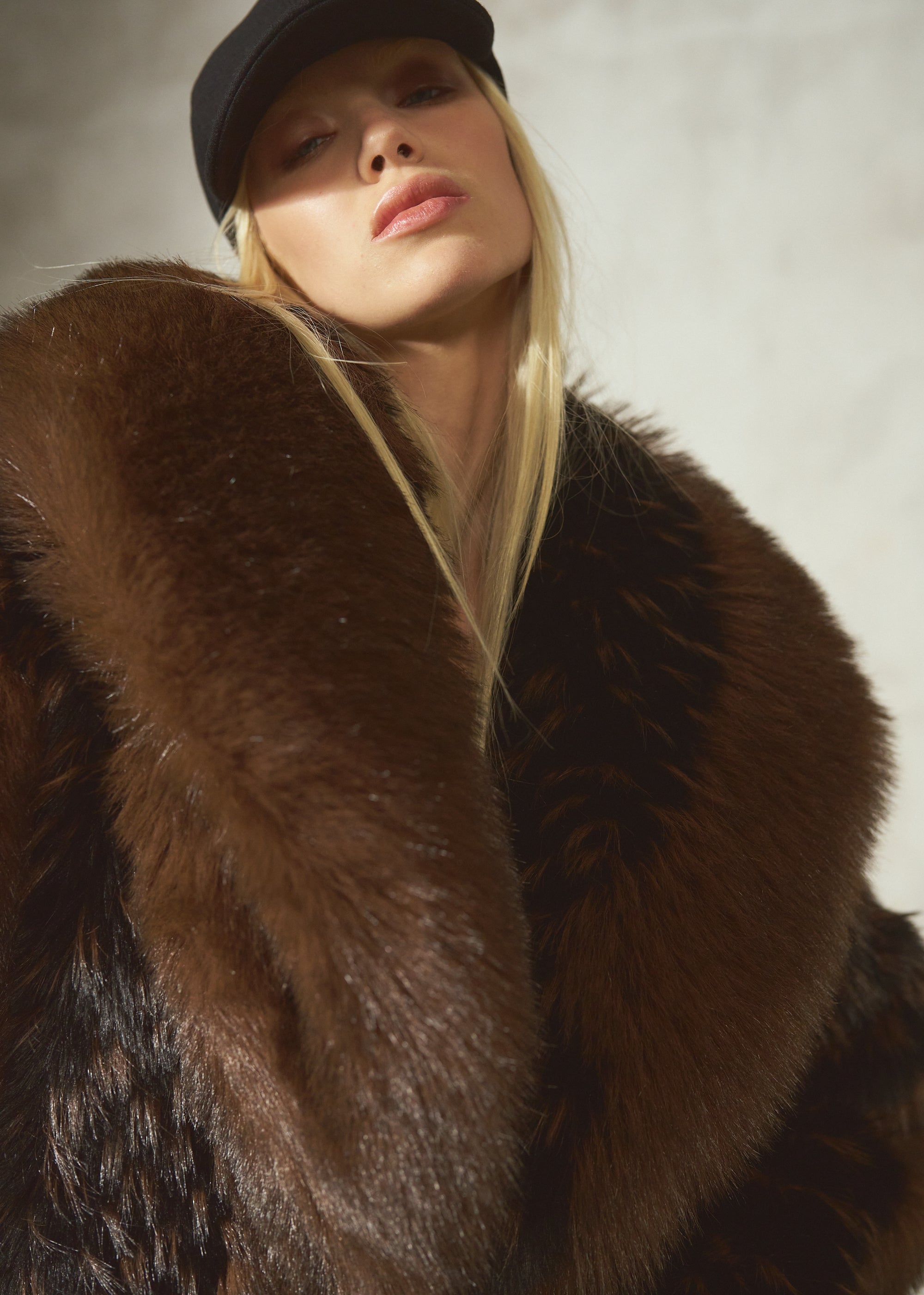 A close-up of a model wearing a luxurious dark brown black fur coat with a wide collar and a black cap, set against a textured light gray background.