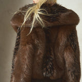 A model wearing a luxurious dark brown black faux 
fur jacket with a large shawl collar, paired with black pants and a black baseball cap, photographed against a textured concrete wall.