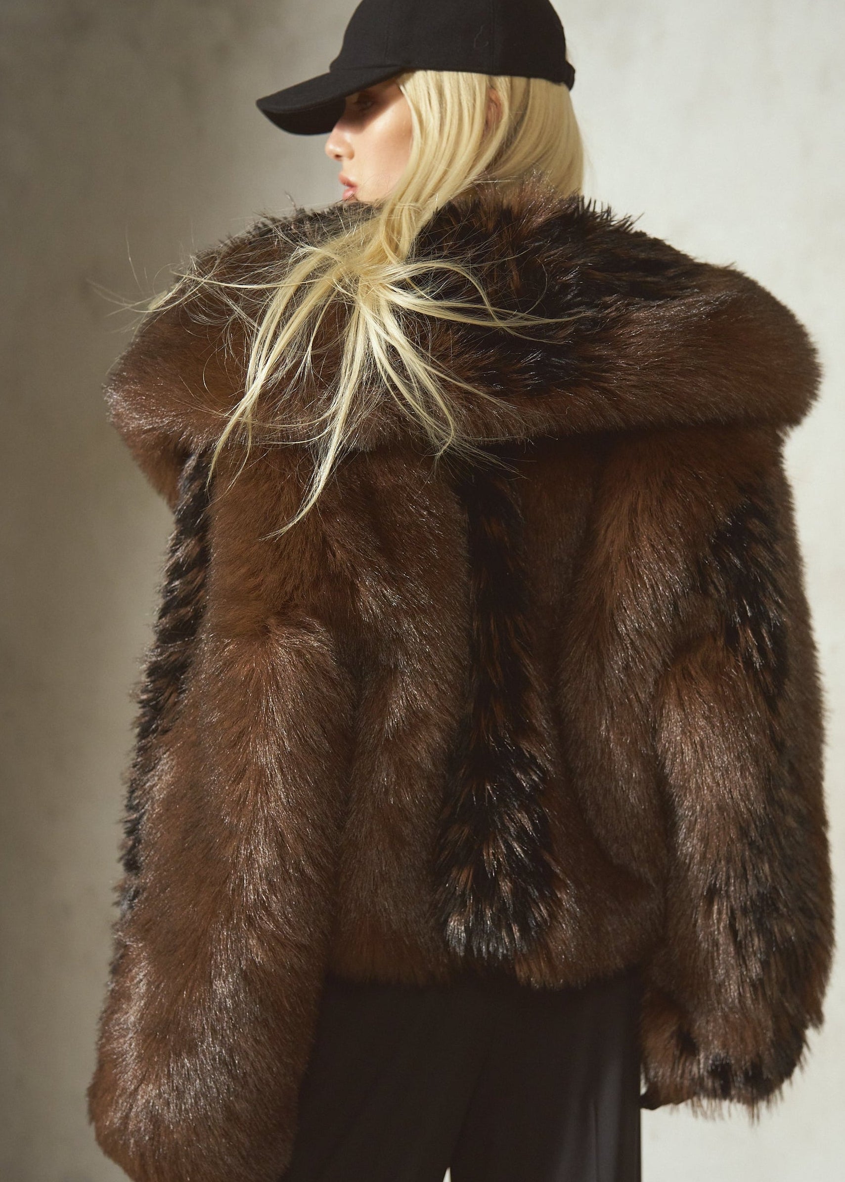A model wearing a luxurious dark brown black faux 
fur jacket with a large shawl collar, paired with black pants and a black baseball cap, photographed against a textured concrete wall.