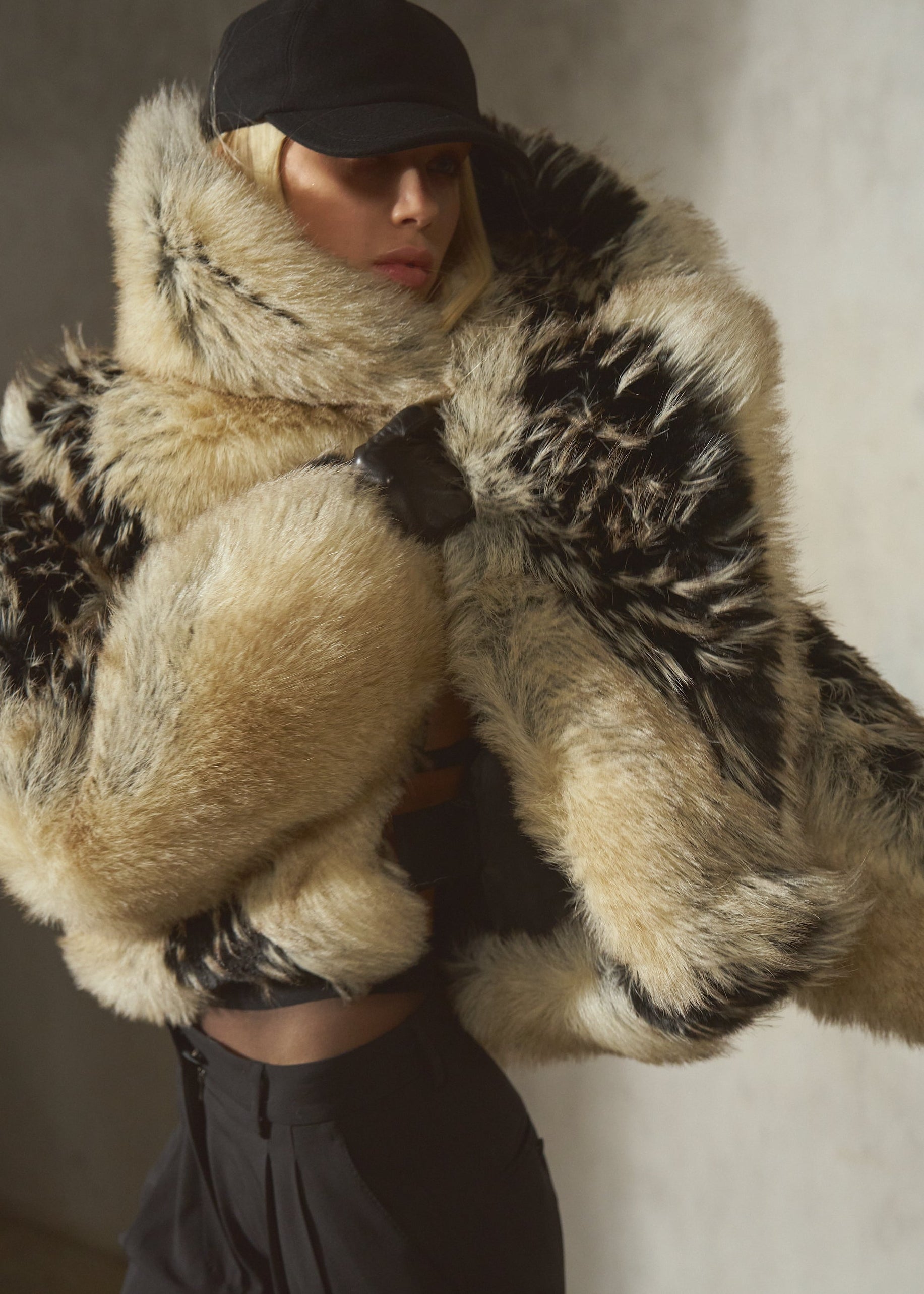 A model wearing a voluminous black and beige faux fur coat with a matching faux fur-lined collar, a black cap, and black pants, posed against a textured gray wall.