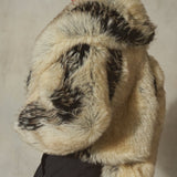 A model wearing a voluminous faux fur jacket with a natural beige and black mottled pattern, styled with a black baseball cap and black trousers against a textured gray wall.