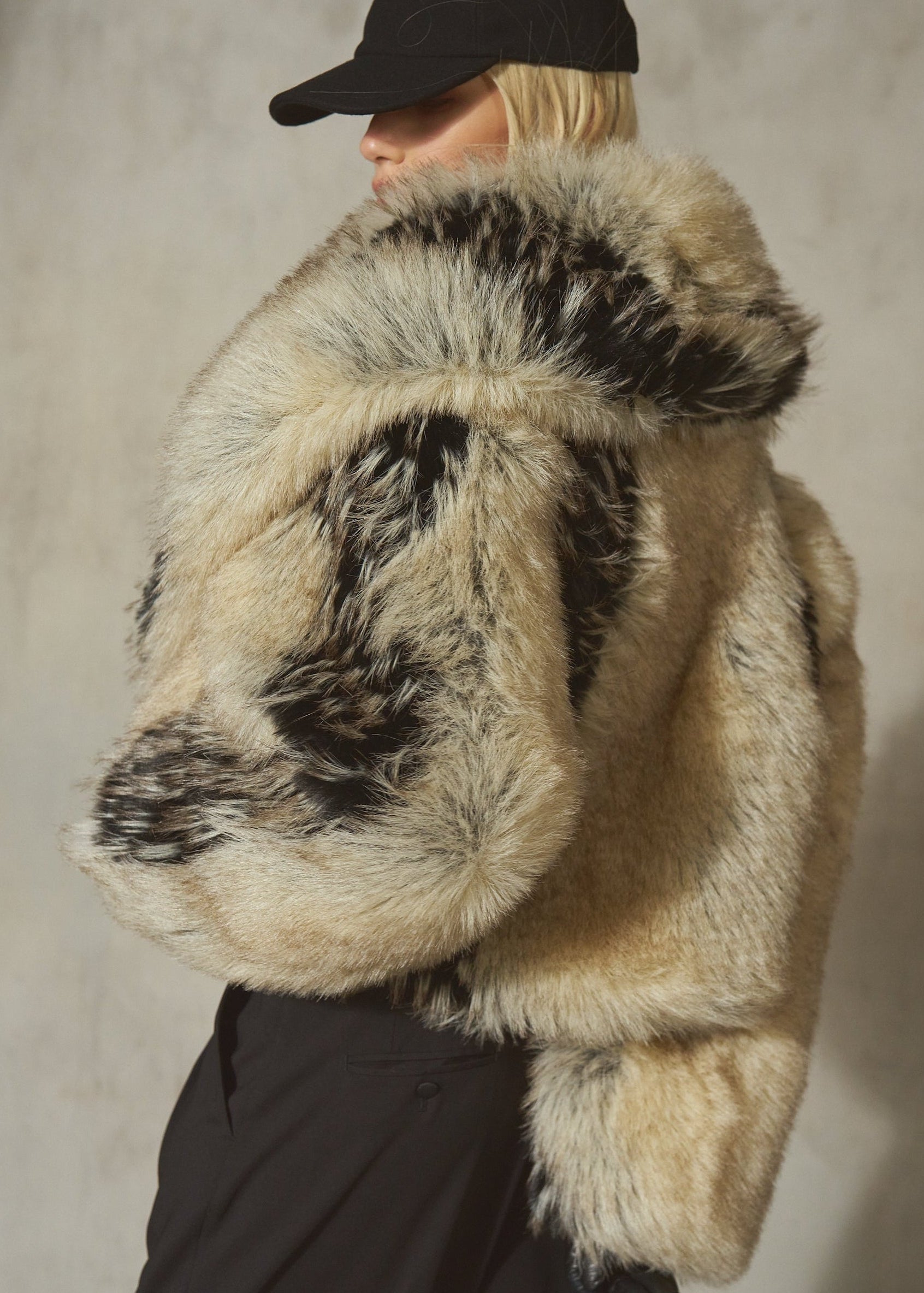 A model wearing a voluminous faux fur jacket with a natural beige and black mottled pattern, styled with a black baseball cap and black trousers against a textured gray wall.