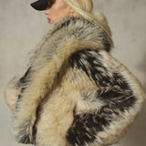 A model wearing a luxurious black and beige fur coat with a black baseball cap, shown from a side profile against a textured gray wall.