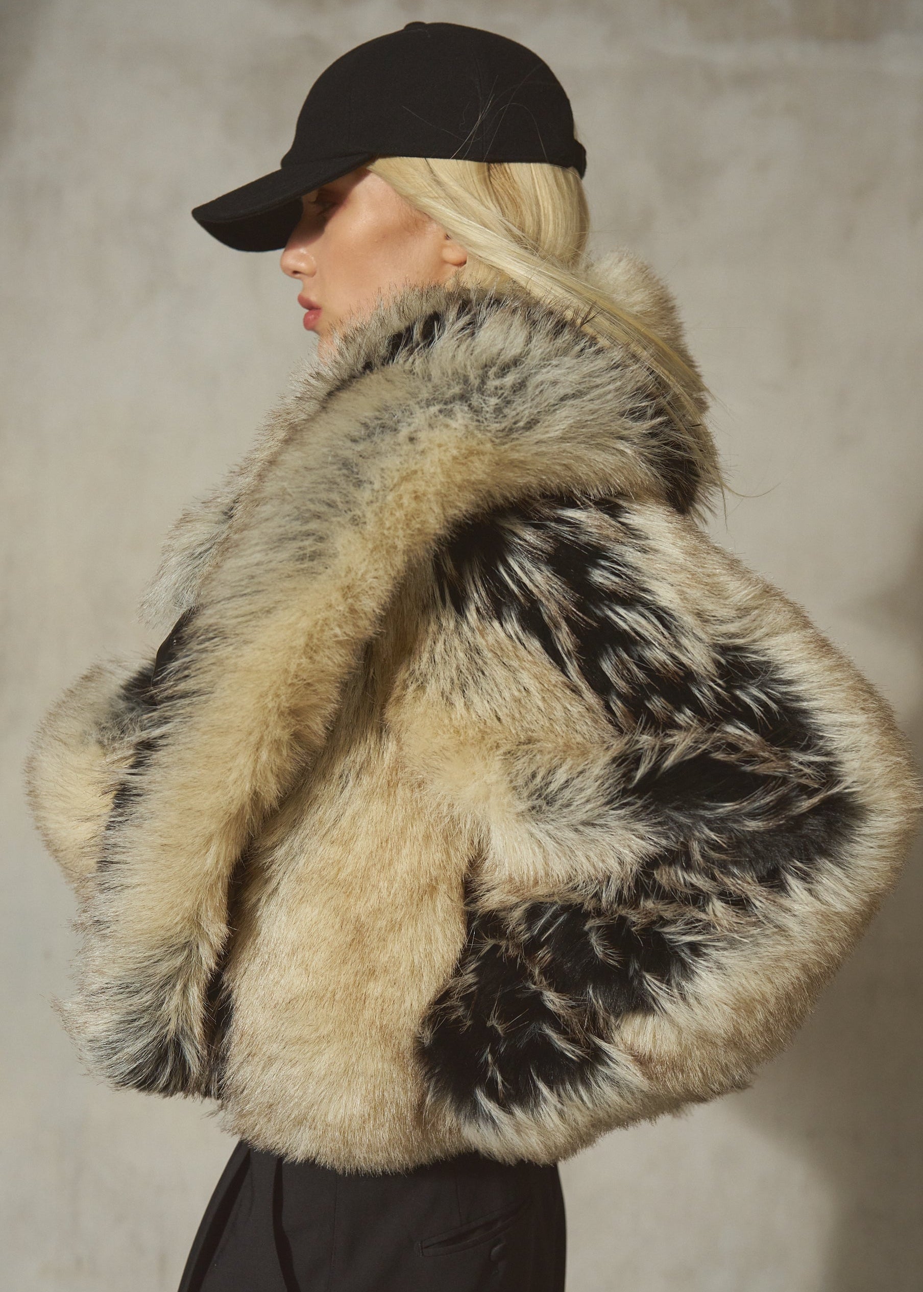 A model wearing a luxurious black and beige fur coat with a black baseball cap, shown from a side profile against a textured gray wall.