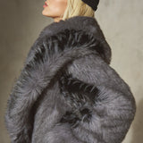 A model wearing a voluminous, oversized dark gray and black faux fur coat with a high collar, paired with a black baseball cap and black trousers, posed against a textured gray studio background.