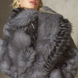 A model wearing a luxurious dark gray faux fur jacket with a large collar, black pants, and a black cap, posed against a textured gray wall.