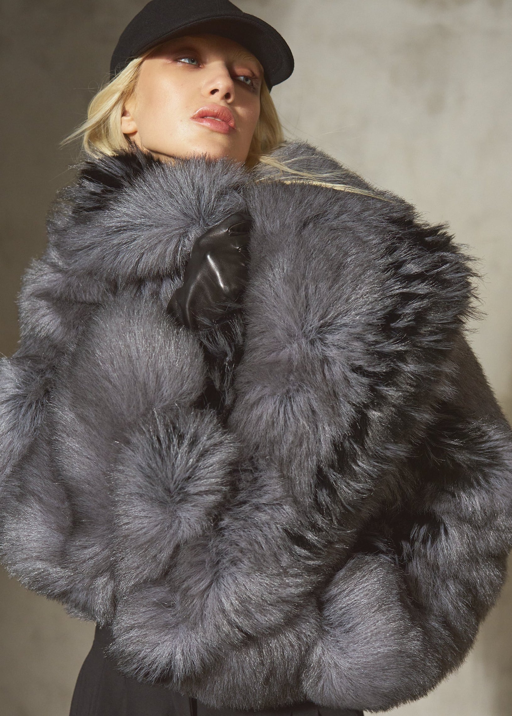 A model wearing a luxurious dark gray faux fur jacket with a large collar, black pants, and a black cap, posed against a textured gray wall.