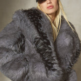 A model wearing a voluminous, oversized dark gray faux fur jacket with a black collar, paired with a black baseball cap and black trousers, set against a textured gray studio background.