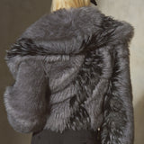 A model from behind wearing a voluminous dark gray faux fur jacket, black trousers, and a black baseball cap against a textured gray wall.