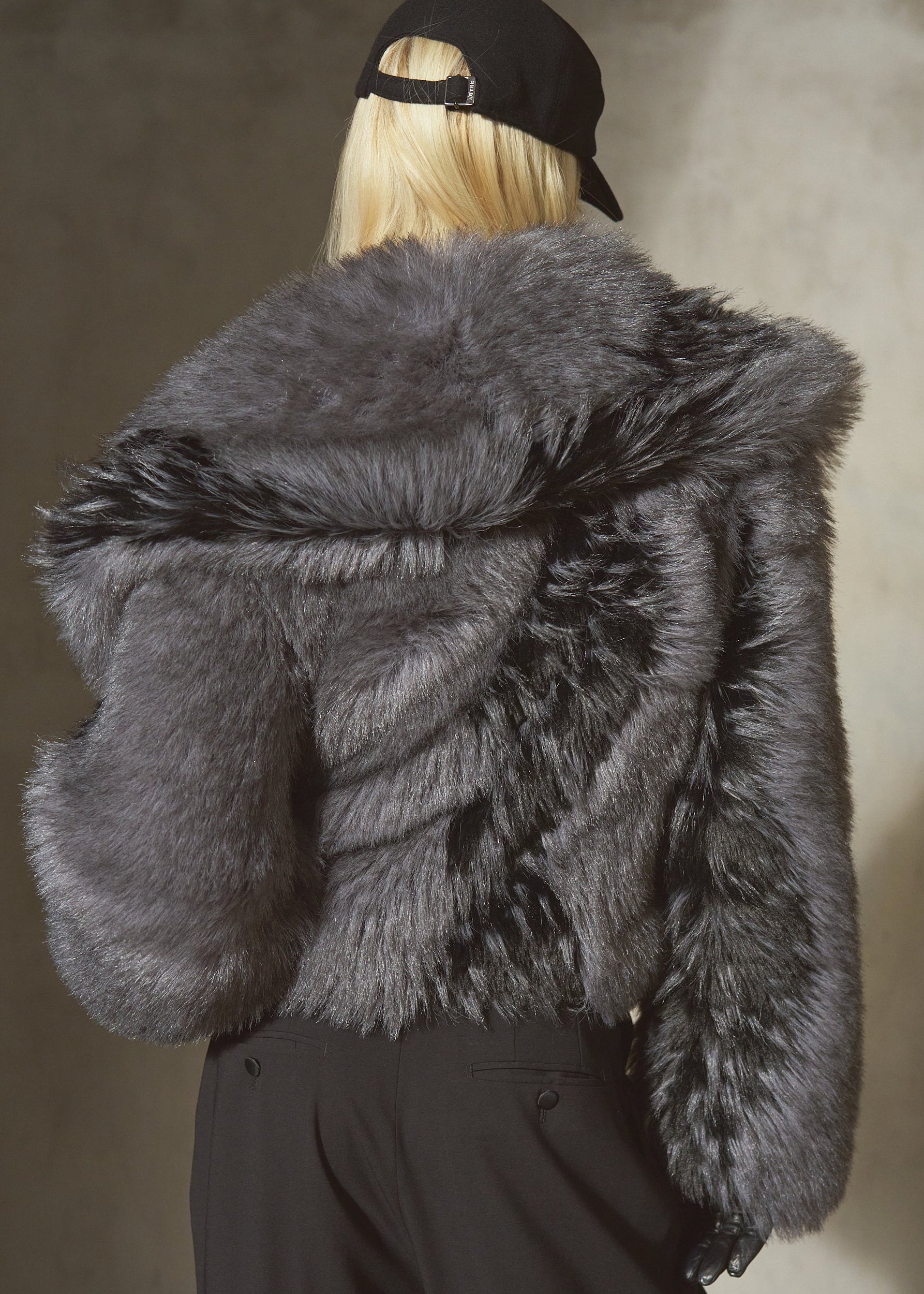 A model from behind wearing a voluminous dark gray faux fur jacket, black trousers, and a black baseball cap against a textured gray wall.