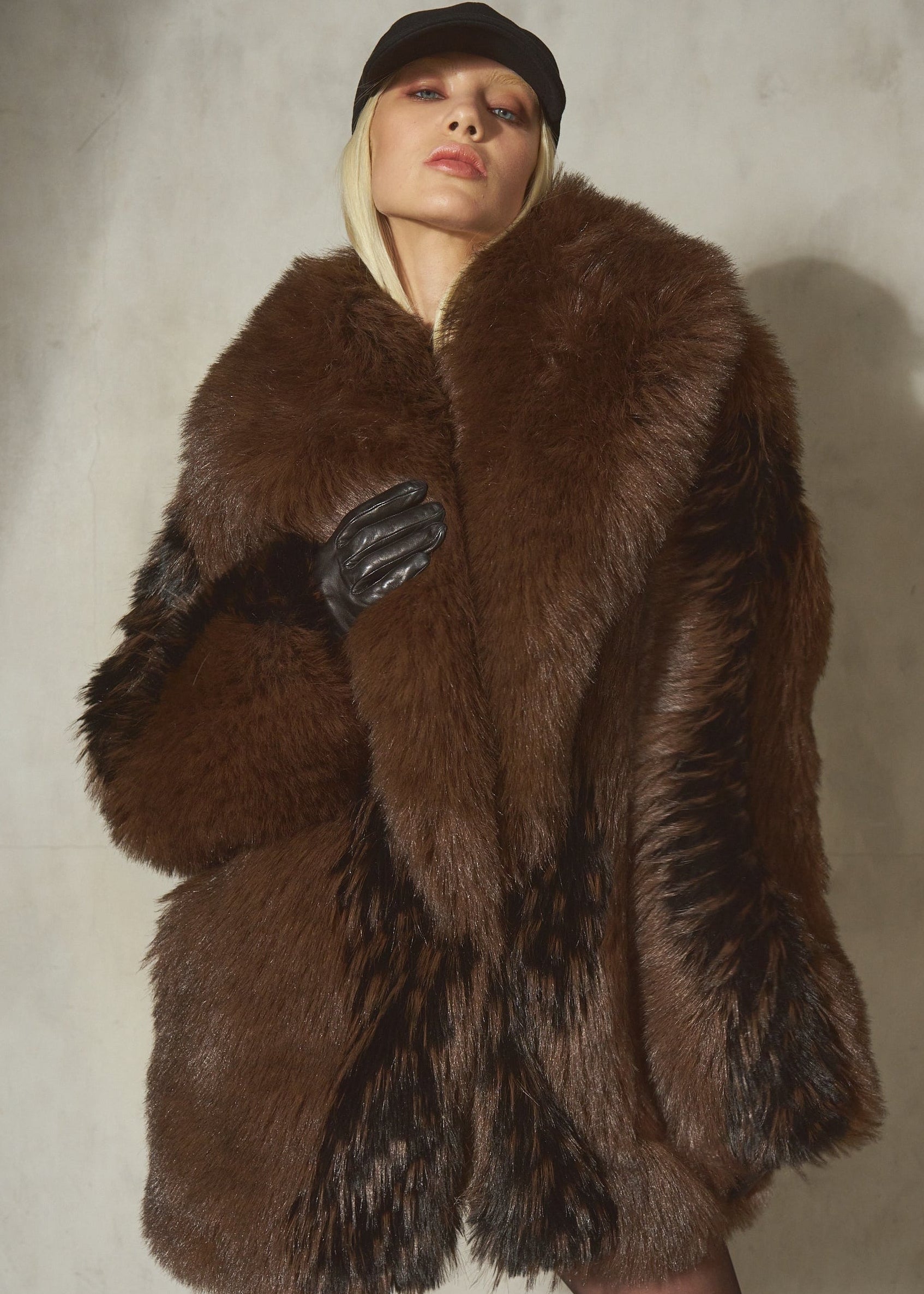 A model wearing a voluminous brown and black faux fur coat with black leather gloves and a black cap, standing against a textured light gray wall.