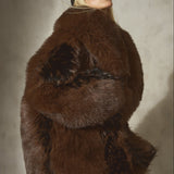 A model wearing a voluminous brown and black faux fur jacket, a black baseball cap, and sheer black tights, standing in front of a textured gray wall.