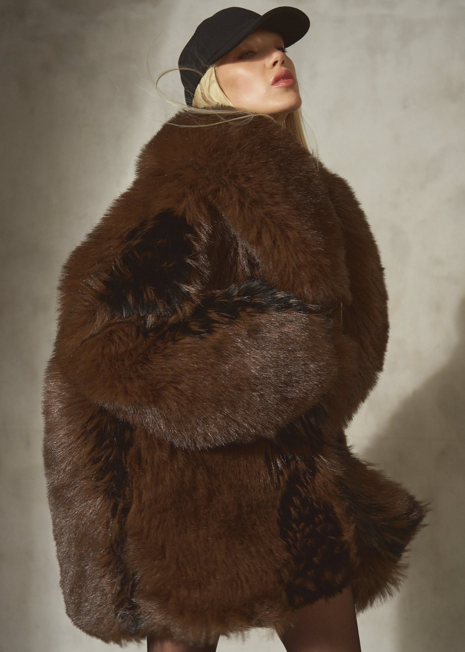 A model wearing a voluminous brown and black faux fur jacket, a black baseball cap, and sheer black tights, standing in front of a textured gray wall.