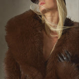 A blonde model wearing a luxurious dark brown fur coat with a wide collar, black leather gloves, and a black wool cap, posing against a textured gray wall.