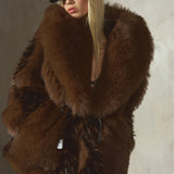 A model wearing a voluminous, oversized dark brown and black faux fur coat with a black wool baseball cap, posed against a textured concrete wall.