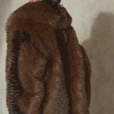 A model wearing a voluminous brown/black faux fur coat with a large shawl collar, paired with a black wool baseball cap and sheer black tights, standing against a textured concrete wall.