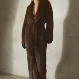 A woman modeling a long brown faux fur coat with a shawl collar and waist tie, paired with a black wool cap, black gloves, and black high heels, standing against a textured concrete wall with dramatic side lighting.