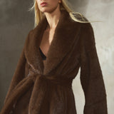 A woman wearing a dark brown faux fur coat with a matching waist tie, a black baseball cap, and black gloves, posed against a textured gray background.