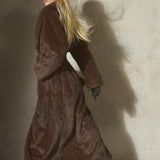 A woman in a long brown faux fur belted coat, black hat, and black gloves walking.
