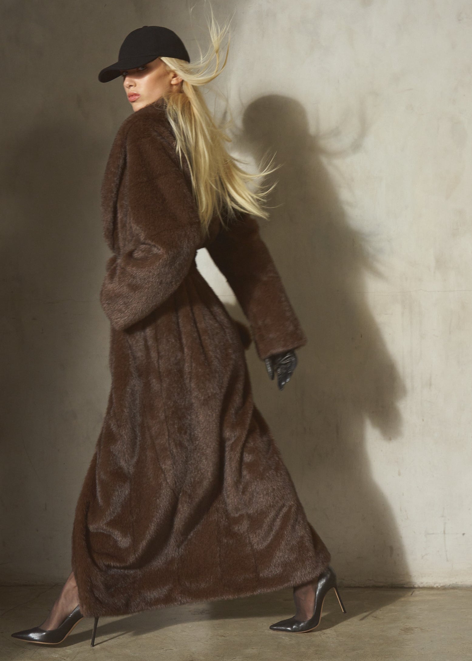 A woman in a long brown faux fur belted coat, black hat, and black gloves walking.