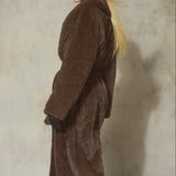 A woman wearing a long brown faux fur coat, black baseball cap, and black gloves, standing against a textured concrete wall.