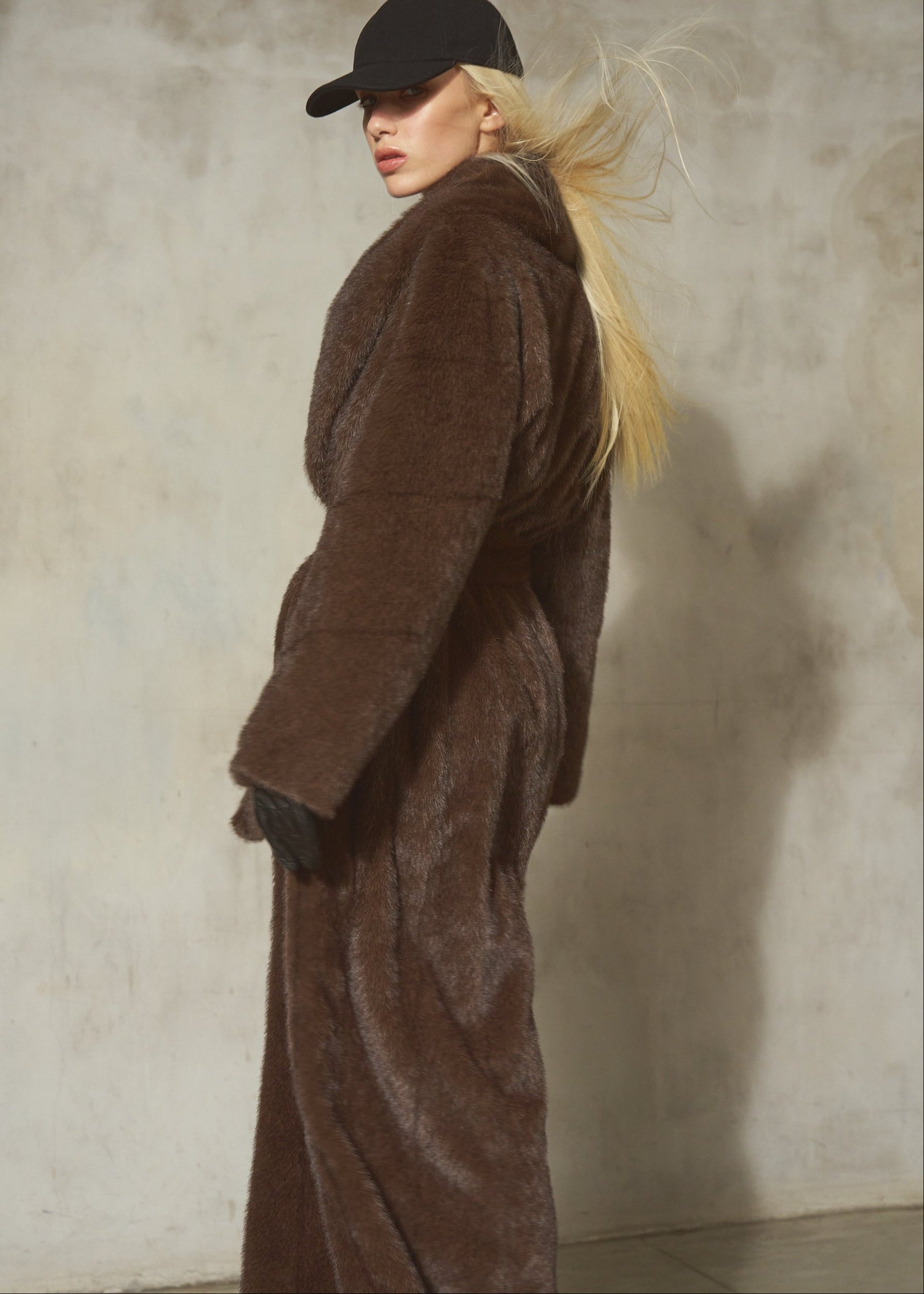 A woman wearing a long brown faux fur coat, black baseball cap, and black gloves, standing against a textured concrete wall.