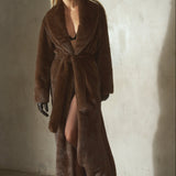 A woman modeling a long, plush brown faux fur coat with a tie waist, black wool cap, and black gloves, walking in front of a textured concrete wall.