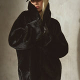 A model wearing a voluminous black faux fur coat, a black baseball cap, and sheer black tights, posing against a textured concrete wall with dramatic side lighting.