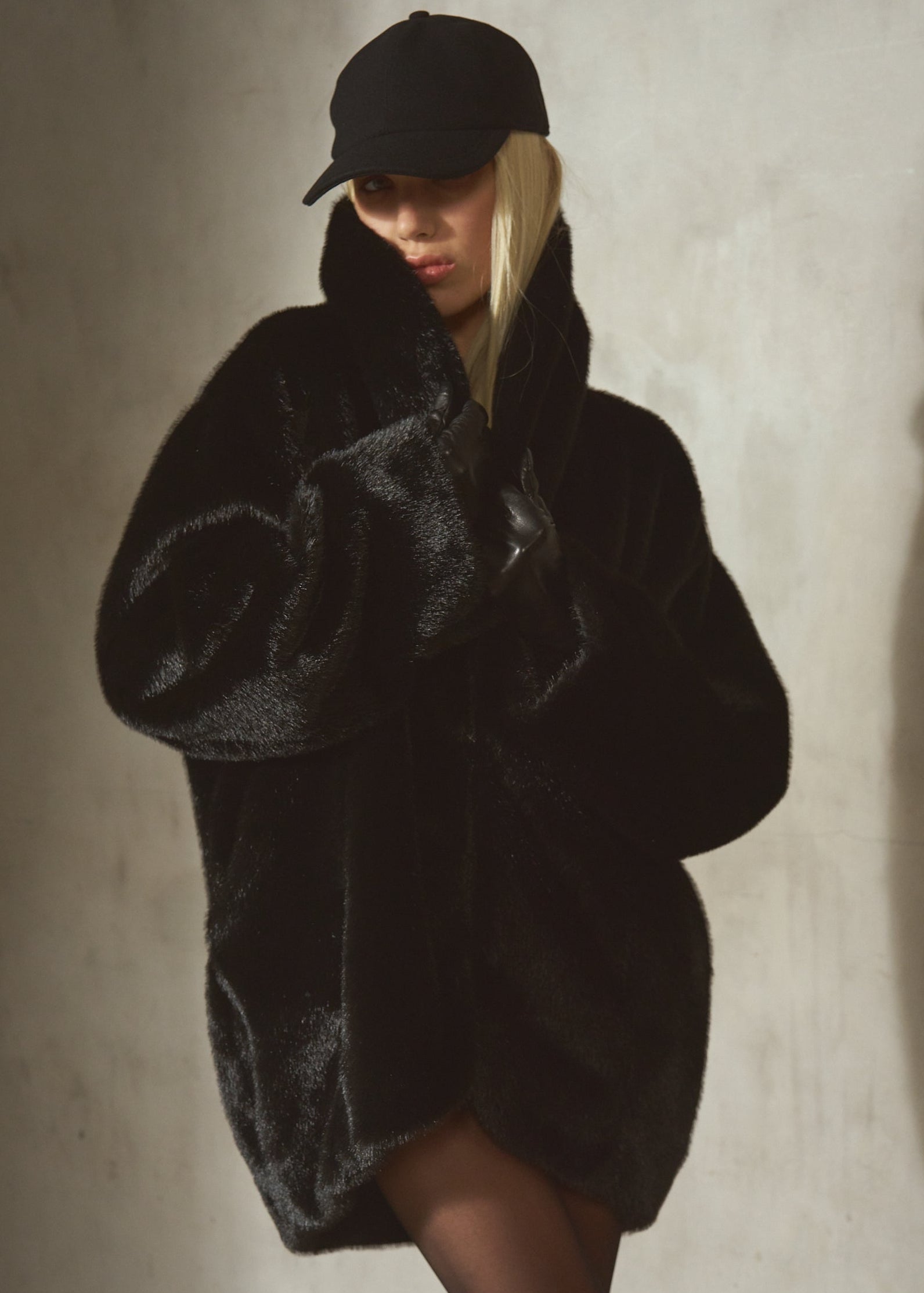 A model wearing a voluminous black faux fur coat, a black baseball cap, and sheer black tights, posing against a textured concrete wall with dramatic side lighting.