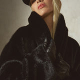 A close-up portrait of a model wearing a luxurious black faux fur coat with a high collar, a black baseball cap, set against a textured neutral background.