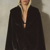 A model wearing a black plush faux fur cocoon jacket and a black baseball cap, standing against a textured gray concrete wall.