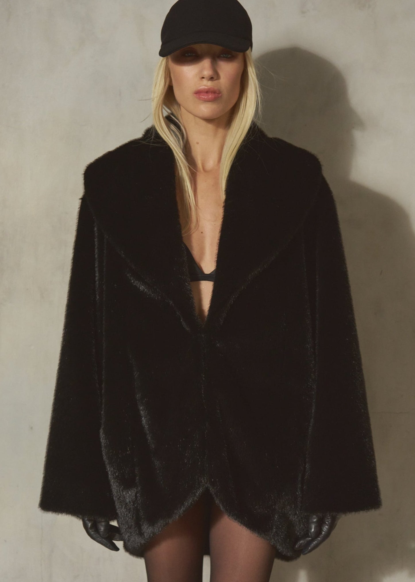 A model wearing a black plush faux fur cocoon jacket and a black baseball cap, standing against a textured gray concrete wall.