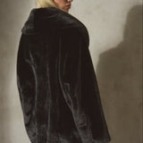 A model wearing a black faux fur coat, black wool baseball cap, and black tights, standing against a textured concrete wall with dramatic side lighting.