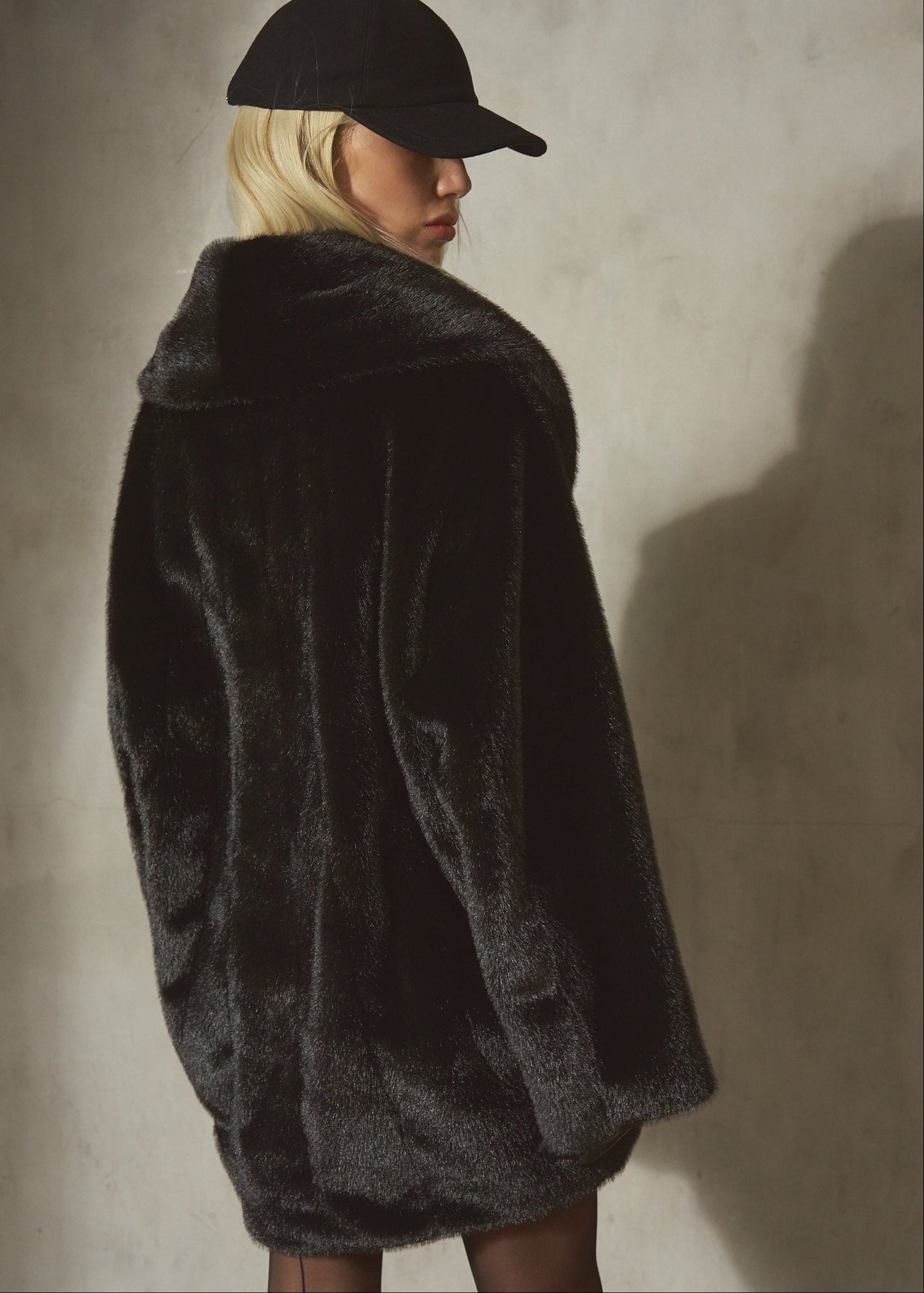 A model wearing a black faux fur coat, black wool baseball cap, and black tights, standing against a textured concrete wall with dramatic side lighting.