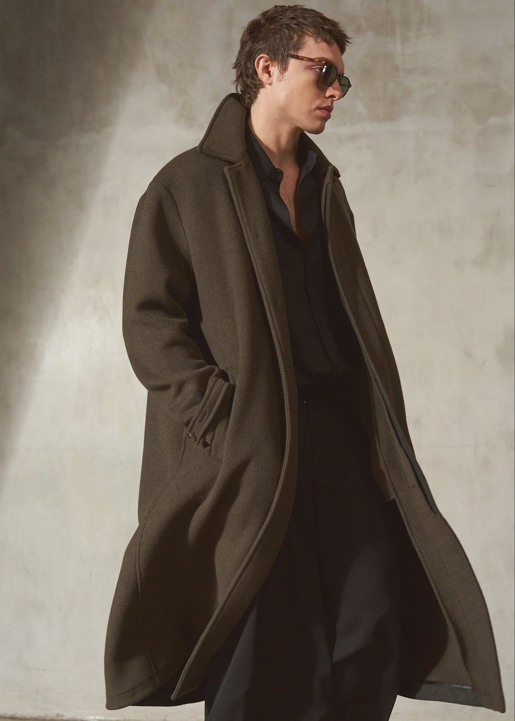 A man wearing a long dark olive-brown overcoat over a black shirt and trousers, standing against a textured concrete wall.