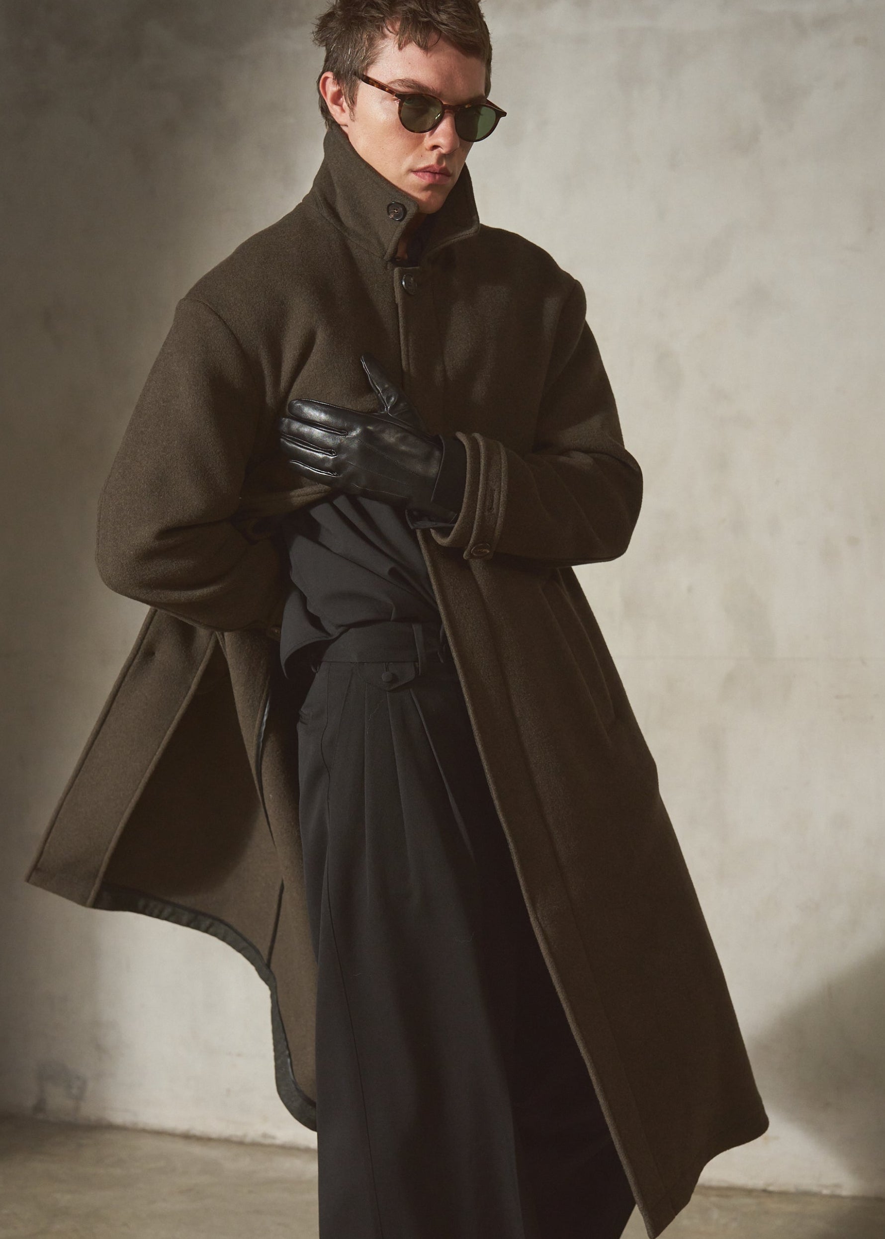 A model wearing a long dark olive green wool-blend coat with black leather gloves and black trousers, standing in a minimalist concrete room.