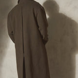 A back shot of a model wearing a long dark olive green coat and dark trousers, casting a long shadow on the wall.