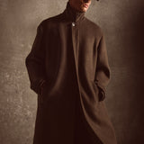 Model wearing a long dark green coat and cap against a textured brown background.