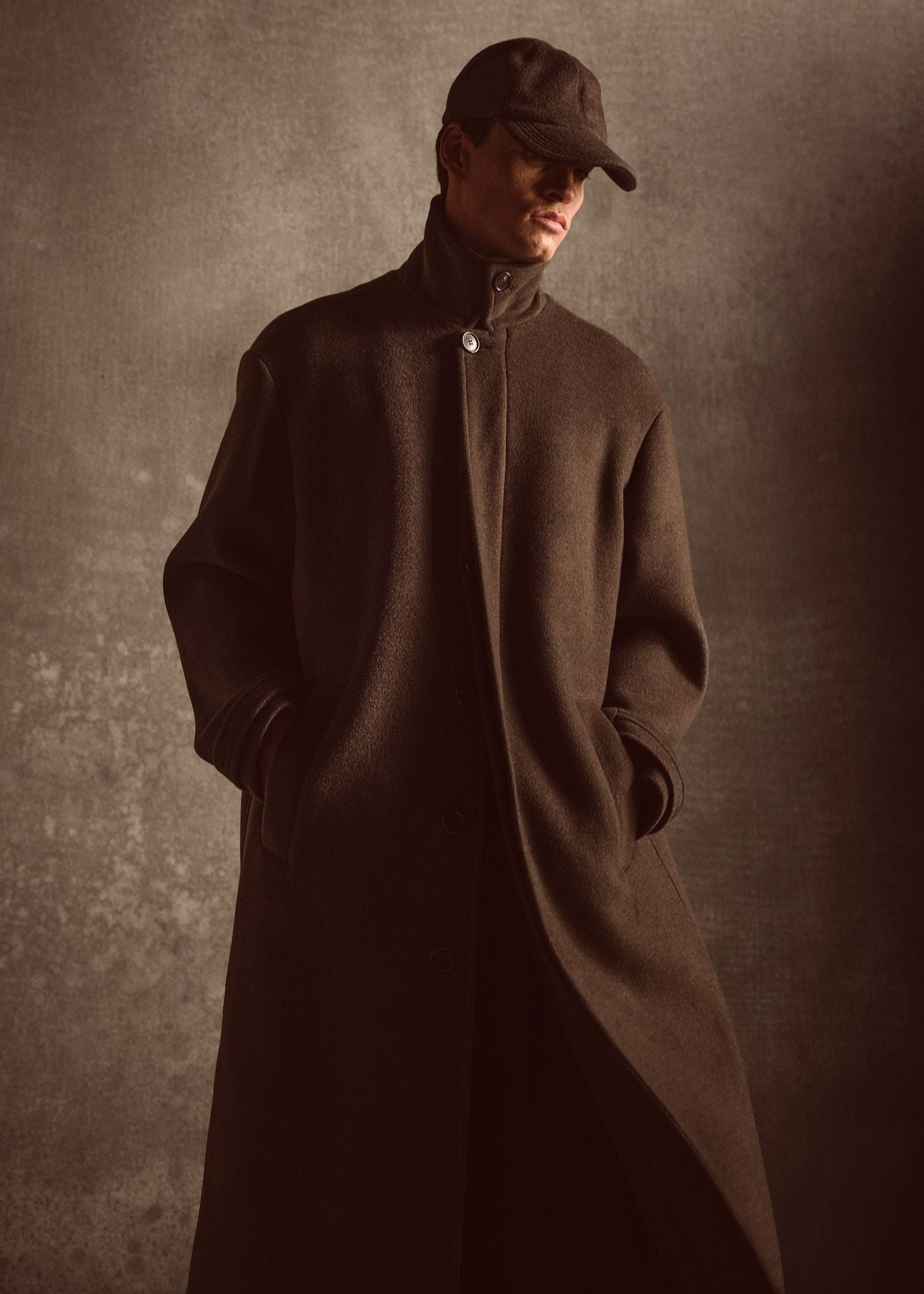 Model wearing a long dark green coat and cap against a textured brown background.